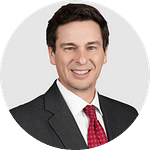 A smiling man with brown hair wearing a dark grey suit, white shirt, and red tie.